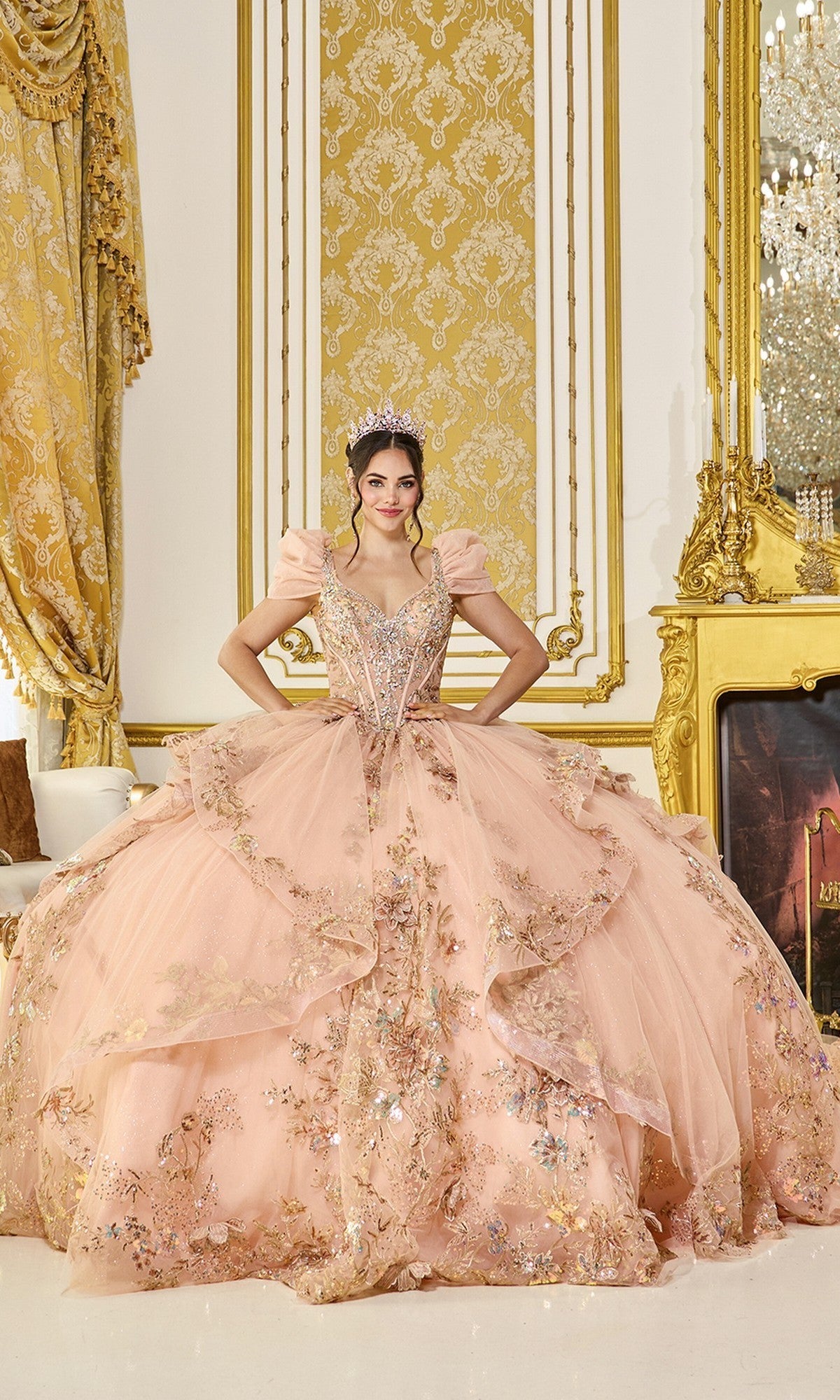 A woman wearing the Quinceanera Dress 5130 By Dancing Queen in rose gold, featuring 3D floral appliques and a sparkling tiara, stands in an opulent room with gold décor, patterned wallpaper, and a decorative fireplace.